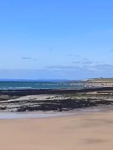 A panoramic view of a beach on a sunny day with a clear blue sky and scattered clouds. The foreground is a sandy beach. Beyond the sand, there are dark, rocky tide pools and then the ocean. In the distance, along the shoreline, there are groups of people walking and standing. The background shows rolling sand dunes and hills.