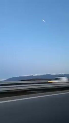 The video shows a landscape viewed from a moving vehicle on a highway. The sky is a clear blue with a few small, light-colored formations. In the distance, there is a mountain range capped with a layer of white, which appears to be snow. In front of the mountains is a brown and green landscape. The highway has a dark gray surface with a white lane marking. There is a metal guardrail visible in the foreground. The camera is inside the moving vehicle.