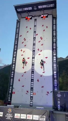 The video shows two climbers ascending a tall, white climbing wall. Both climbers are wearing helmets and climbing gear. The climber on the left wears a red shirt and white pants, while the climber on the right wears a black shirt and black pants. Red climbing holds are scattered across the wall. Digital timers are mounted at the top of the wall, displaying the climbers' times. The wall is brightly lit, and the sky above is a clear blue. Mountains are visible in the background. The scene appears to be an outdoor competition.