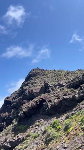 The video shows two people standing on top of a rocky hill under a blue sky with scattered clouds. The hill is covered with dark-colored rocks and some green vegetation. One person is standing and looking around, while the other is holding a phone or camera and appears to be taking a picture.