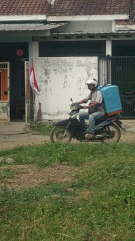 A person wearing a white helmet and a gray shirt rides a dark-colored motorcycle with a large blue box attached to the back. The motorcycle is parked on a dirt road next to a grassy area and some trees. In the background, there is a house with a red and orange striped door and a white wall. An Indonesian flag is visible hanging on the house. The scene appears to be outdoors during the daytime.