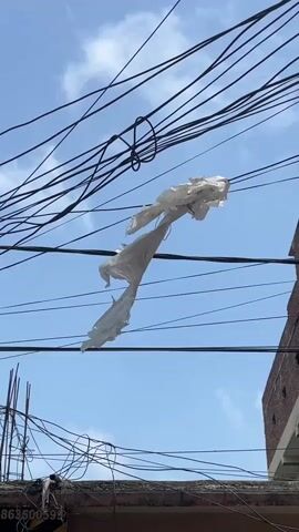 A tattered white plastic bag is caught on overhead wires against a bright blue sky with some wispy clouds. The plastic is ripped and flapping in the wind. Below the wires, the edge of a building with brickwork and some visible rebar is present. Another set of tangled wires is visible closer to the building.