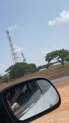 The video shows two communications towers against a sky with scattered clouds. The towers, made of metal lattice, are partially obscured by treetops. The camera, positioned inside a moving vehicle, offers a view through the car window and side mirror. The vehicle is presumably traveling on a road in a rural or suburban area. The scene is bright and appears to be during the daytime.
