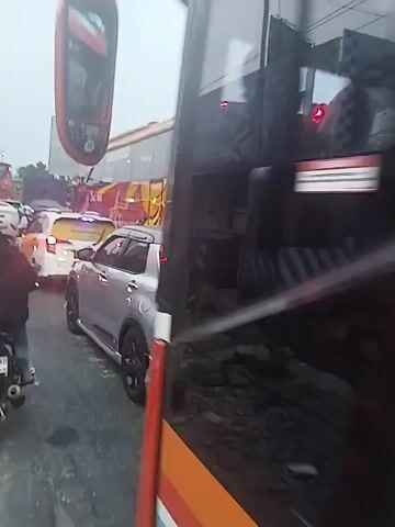 The video shows a busy road with heavy traffic, including buses, cars, and motorcycles. The buses are large and brightly colored, with the words "JETBUS PARIWISATA" and "BUS PARIWISATA" visible on their sides.  The cars are mostly silver or white. Many people on motorcycles are moving through the traffic. The sky is overcast. A building with signs is visible on the side of the road.  The road appears congested.