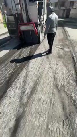 The video shows a street being paved with asphalt. A paving machine is laying down a layer of asphalt, leaving tire tracks in the fresh material. A man in a gray hoodie walks alongside the machine. In the distance, a small yellow bulldozer-like vehicle is also present. Buildings line the street, and the sky is clear and sunny. The camera moves, showing different angles of the paving process and the surrounding environment.