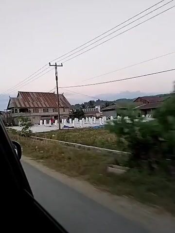 The video captures a view from a moving vehicle, likely a car, showing a rural or suburban landscape. The foreground shows a road and the side of the vehicle. In the background, there are buildings, a large house with a distinctive tiered roof, and an area with many white, tarp-covered objects arranged in rows, possibly for drying or storage. There are also some trees and utility poles with overhead wires. The sky is overcast. The video appears to be a casual recording of the scenery passing by.