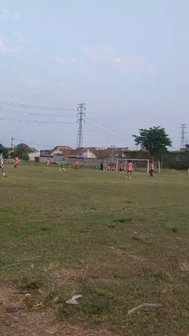 A group of people in soccer uniforms are playing soccer on a grassy field. There are two goalposts visible. In the background, there are houses, trees, and power lines. The sky is clear and the sun is shining.