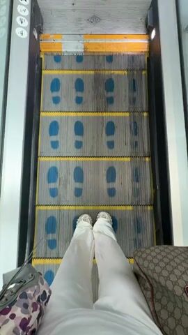 The video shows a first-person perspective of someone riding an escalator. The person is wearing white pants and shoes. They have two bags: one with a floral pattern and another with a Gucci logo. The escalator has blue footprints painted on the steps. The person reaches the top of the escalator and steps onto a white tiled floor. The floor transitions into a gray carpet.