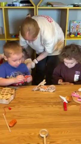 Three young children and an adult woman are at a wooden table. The children are playing with wooden toys and the woman is assisting them. The boy on the left wears a blue shirt and is putting together wooden pieces. To his right, a boy in a maroon shirt is also playing with wooden toys. Behind them is a young girl in a white shirt, standing and also playing with toys. The adult woman is wearing a white shirt with pink writing on the front. The table is covered with a variety of wooden toys, including pieces that resemble food, tools, and puzzle-like shapes. A yellow bookshelf is in the background, filled with toys and books.