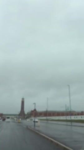 A video captures a view from a moving vehicle on a wet road under a cloudy sky. On the left side of the road, a tall, slender, reddish-brown tower with a white top stands out against the overcast sky. To the right of the tower, a long, red-brick building with white-trimmed windows stretches horizontally. The road is lined with streetlights. Several cars are visible on the road, some moving and some stationary. Traffic signals and signs are present. The road surface appears wet and reflective, indicating recent rain. The overall atmosphere is overcast and somewhat gloomy.