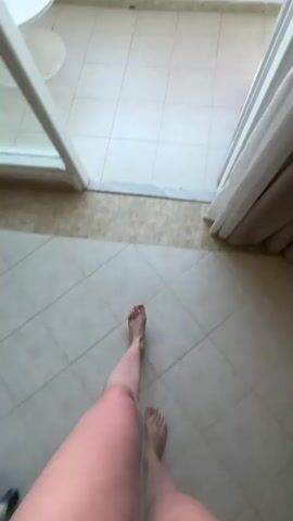 The video shows a view from a balcony overlooking a tropical resort. Initially, a person's lower legs and feet are visible as they walk onto the balcony. The camera pans up to show the balcony, which has a white table and white latticework railings. A pink and orange towel hangs over the railing. Beyond the balcony, there is a view of palm trees, swimming pools, and buildings of the resort. The sea is visible in the distance under a clear blue sky.