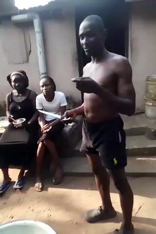 Outside a modest dwelling, a shirtless man in black shorts stands before two women seated on a concrete block. He holds a plate and a knife, sampling its contents. One woman, in a black dress, gives a peace sign. The other, in a white shirt, looks down. Several large pots of liquid sit on the ground. The camera then shifts focus to another man, seated in a small, shaded structure. He is wearing a pink shirt, and sampling liquid from a plate as well, amid pots of liquid.