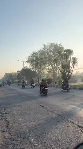 The video shows a street scene during sunrise. Several people are riding motorcycles on a paved road. Trees and a gas station are visible in the background. The sun is low in the sky, casting long shadows. The overall atmosphere is peaceful and calm.