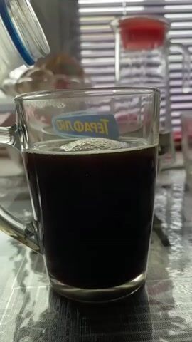 A close-up shot shows milk being poured into a clear glass mug filled with a dark liquid, likely coffee. The milk creates a frothy layer on top of the coffee. The mug has a handle and a logo on the side. The background is blurred but suggests an indoor setting with blinds, a glass pitcher with a red lid, and a bowl of food. The surface beneath the mug appears to be a patterned placemat. The video captures the pouring process from start to finish, showing the milk stream and the resulting change in the drink's appearance.
