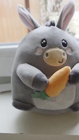 A gray stuffed donkey toy sits on a white surface. The donkey has large eyes, rosy cheeks, a tan snout, and a small smile. It holds an orange plush carrot with green leaves in its paws.  A red tag with "LEGAMI" printed in white is attached to the donkey's side. A small card with an illustration of the same donkey is visible next to the toy. Faintly in the background, a blurry window pane is seen. Music with male vocals plays in the background.