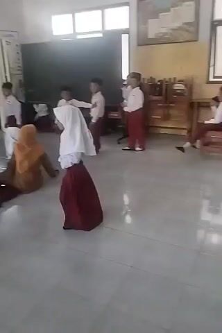 The video shows a group of children in a room, likely a classroom. Most of the children are wearing white shirts and maroon skirts or pants. Some children are standing, while others are sitting or lying on the floor. One child in the foreground is wearing a white headscarf. In the background, there is a blackboard, windows, and some furniture. The children appear to be engaged in some activity, possibly a game or a performance. The overall atmosphere seems casual and playful.