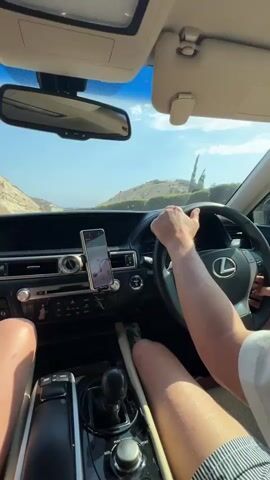The video shows the interior of a Lexus car from the perspective of a passenger. A person's hands are on the steering wheel, driving the car on a paved road. The passenger's legs are visible in the foreground, resting in the car. The dashboard features a smartphone mounted on a holder, displaying a navigation app. The exterior view through the windshield reveals a sunny day with a landscape of dry, hilly terrain and a bright blue sky. The car appears to be moving along a coastal road, with glimpses of the sea in the distance. The overall scene depicts a road trip or travel.