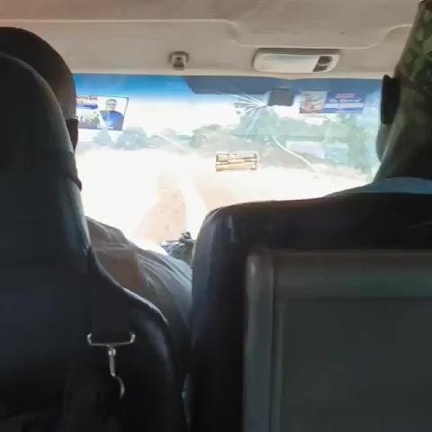 The video shows a view from the back seat of a vehicle traveling down a dirt road. Two people are seated in front, partially visible from the back. The person on the right is wearing a patterned hat and a light blue shirt. The person on the left has a black bag with a silver clip hanging from the seat. The vehicle's windshield is cracked. There are two small posters or stickers on the windshield. The road ahead is dusty and unpaved, with some trees and greenery visible in the distance. The lighting is bright, indicating it is daytime. The vehicle appears to be moving.