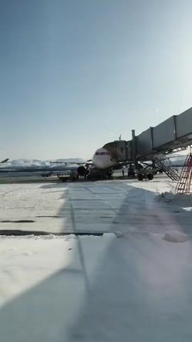 The video shows a passenger airplane parked at an airport gate on a snowy day. The plane is white with grey accents, and the front of the plane is connected to a jet bridge. The ground is covered in snow, and there are mountains visible in the distance. The sky is blue and clear. There are ground crew workers visible around the plane. A luggage cart is near the plane.
