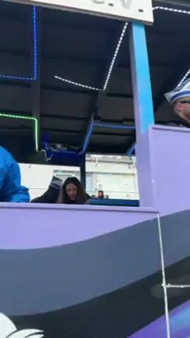 A man in a blue jacket and a white and blue hat sings into a microphone atop a carnival float.  Another person with long dark hair sits behind him.  The float is painted with a white, blue, and purple design including the word "PRINZENPAAR".  The camera pans to reveal more of the float and the surrounding environment, including a green tractor, a person in a yellow traffic vest, and people in costume.