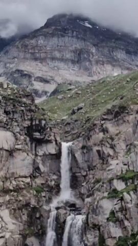 A cascading waterfall plunges down a rugged, rocky mountainside. The water flows in multiple streams over the rough terrain, creating a dynamic and powerful visual. The surrounding rocks are layered and appear weathered, with patches of green vegetation clinging to the slopes in some areas. The top of the mountain is shrouded in clouds, adding to the dramatic and remote atmosphere of the scene.