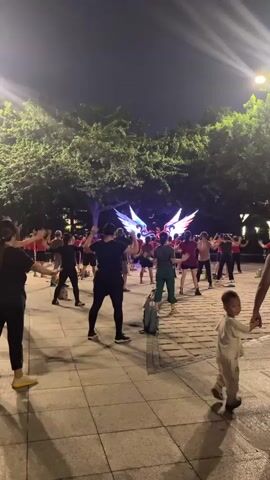 A group of people are exercising outdoors at night in a paved area with trees and lights in the background. There is a bright, colorful light display with what appears to be large wings in the distance. The individuals are dressed in casual clothing and are performing synchronized movements, suggesting a group fitness class or dance. One person in the foreground is facing away from the camera and appears to be demonstrating exercises. A young child is also visible in the lower right corner of the frame, looking towards the camera. The lighting is dim, but the scene is illuminated by artificial lights from streetlamps and the distant light display.