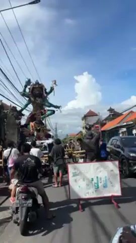 Here is a description of the video:
A video shows a religious parade or procession moving down a street. The focus of the parade is a large, brightly colored statue of a multi-armed deity, with a green body and ornate decorations. The statue appears to be carried on a platform by a group of people. The street is busy with onlookers, some of whom are taking pictures. The sky is blue with scattered clouds. Power lines are visible above the statue. There are buildings with traditional architectural styles in the background.