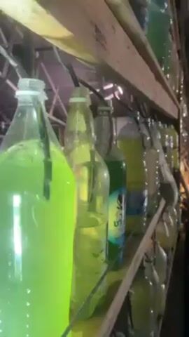 A close-up shot shows a series of plastic and glass bottles filled with green and yellow liquids on wooden shelves. Hoses are connected to the bottles. One bottle has a label with writing in Hebrew and the image of an alien. The bottles are various shapes and sizes.  The overall impression is a homemade setup.