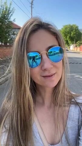 A woman wearing reflective sunglasses and a gray top is filming herself on a sunny street. Her long blonde hair is blowing in the wind. She is speaking, smiling, and gesturing with her hands. The street is paved and has houses and trees visible in the background. There are utility poles and lines overhead. The reflection in her sunglasses shows the street and buildings behind her.