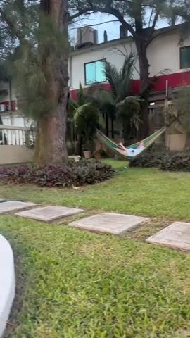 The camera pans from a hammock to a small swimming pool in a yard. A person is lying in the hammock, which is strung between two trees. The yard has a lush green lawn and stepping stones leading to a gate that opens onto a beach. A glass of red wine sits on the edge of the pool. The sky is overcast. The camera movement is smooth and deliberate.