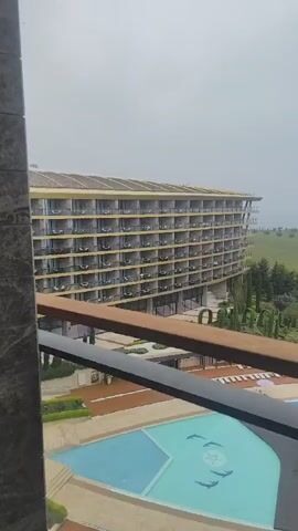 The video shows a view from a balcony, panning from left to right. A large, multi-story hotel building with many balconies dominates the left side of the frame. Below, there are swimming pools with turquoise water, surrounded by landscaped areas and lounge chairs. As the view pans right, a large pool with a gazebo and a pattern on the bottom comes into view. Further to the right, lush green hills and trees stretch towards a body of water. In the distance, a ship is visible on the water, under a cloudy sky. The balcony railing is in the foreground, slightly obscuring the view. A few people can be seen near the pools. The overall impression is of a high-end resort on a cloudy day.