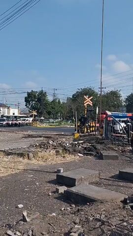 A train passes through a railroad crossing. The train is red and blue and has "KAI" written on it. The crossing has a yellow X-shaped sign. The area is dusty and cluttered with debris. The sky is clear and blue.
