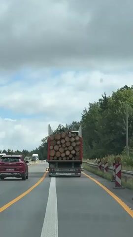 A truck carrying a large load of logs is driving on a highway. The truck has a German license plate that reads MAB FB 631. There are other vehicles on the highway, including a red car and a white van. The sky is cloudy with patches of blue sky. There are trees on the side of the road. A speed limit sign of 80 is visible. Another sign reads "Burghausen 100km." The truck appears to be driving in the right lane. The logs are stacked high in the truck bed.
