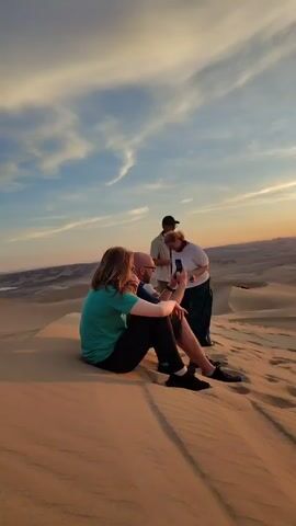 The video depicts a group of people on a sand dune landscape, presumably at sunset. A group of four individuals is seen initially, with three standing and one sitting on the sand. The person sitting is interacting with one of the standing individuals, possibly showing something on a mobile phone. The other two stand nearby, observing. 

As the camera pans slightly, a red off-road vehicle with people in it is visible. Two young women then walk towards the camera, with one briefly striking a pose. The scene is set against a backdrop of rolling sand dunes and a sky painted with colors of the setting sun, suggesting a desert environment. A yellow off-road vehicle is also visible in the distance.
