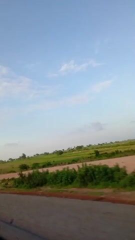 The video shows a view from a moving vehicle traveling along a paved road. To the right of the road, there is an expanse of green fields with scattered trees and some buildings in the distance. The sky is blue with a few scattered white clouds. The terrain appears to be gently sloping. The lighting suggests it is daytime, possibly late afternoon or early morning given the soft light. No people or animals are clearly visible in the frame.