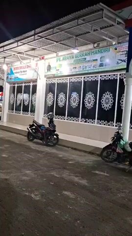 The video shows a stationary wide shot of a gas station at night. The gas station has a white and red canopy over the pumps, which are visible in the background. In the foreground, there is a building with a white facade and a black roof. The building has several windows with decorative grilles and is lit by overhead lights. Two motorcycles are parked in front of the building. The ground is paved and appears to be an asphalt or concrete surface. The sky is dark, indicating it is nighttime.