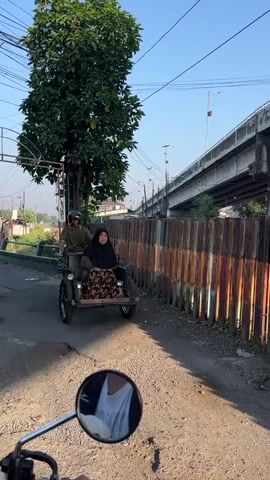 A motorcycle is being ridden on a dirt road. The rider is facing away from the camera, and a woman is sitting in a sidecar attached to the motorcycle. She is wearing a hijab and a patterned dress. They are moving away from the camera. The environment appears to be a rural or semi-urban area, with a large tree on the left and a rusted metal fence on the right. An elevated bridge or overpass is visible in the background. The sky is clear and blue, and the sun is shining. The camera's perspective suggests the video is being filmed by another motorcyclist.