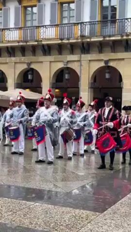 Certainly! Here's a concise, forensic description of the video:

**Scene:**

The video features a group of approximately 12 individuals in a city square.

**People:**

All are adult humans, presumably men, dressed in what appears to be historical military or parade uniforms. The uniforms include ornate jackets, white pants, hats with feathers, and distinct color variations depending on rank or role.

**Objects:**

Each person holds a drum, adorned with decorative cloth bearing distinctive insignia and text. The drums are actively played. The square has a wet, reflective stone-paved surface. The background shows buildings with archways and multi-storied facades with shuttered windows.

**Actions:**

The individuals are performing as a drumming ensemble. They are playing their drums in coordinated rhythm. The drums make a deep percussive sound.

**Interactions:**

The ensemble plays in unison.

**Environment:**

The scene is outdoors in a European-style city square. It appears to be after a rain, as indicated by the wet pavement.