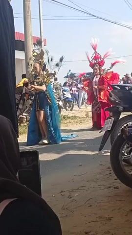 The video shows an outdoor event with several people. In the foreground, a person wearing a black headscarf and dark clothing is partially visible, holding a smartphone. In the middle ground, two individuals are dressed in elaborate costumes. The person on the left wears a dark blue and gold outfit with a long blue cape and a headdress adorned with peacock feathers. The person on the right wears a bright red, elaborate gown with a tall, feathered headdress. They are standing on a dirt road lined with buildings and parked motorcycles. In the background, more people are visible, some on motorcycles, and a man in a blue t-shirt stands near the person in the red costume. The sky is clear and blue. The event appears to be a parade or festival.