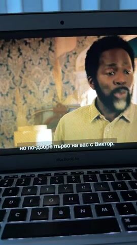 A close-up shot shows a laptop screen displaying a scene from a movie. The screen is a MacBook Air. The scene features two men. One man, who is Black with a beard and graying temples, is in the foreground, looking to the right and speaking. Subtitles in Bulgarian read "но по-добре първо на вас с Виктор." In the background, a white man with graying hair and a beard is visible, looking intently forward and speaking. The subtitles for his dialogue read "Искам да поговоря с двама ви." Further in the background, another Black man is dimly visible. The environment appears to be indoors, possibly a house, with patterned wallpaper and shelves of items in the background. Below the movie screen, the keyboard of the MacBook Air is visible, with its keys illuminated.