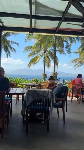 A camera pans across an outdoor seating area of a restaurant. Several wooden tables and chairs are visible, some with jackets draped over them. In the background, palm trees frame a view of the ocean and distant mountains under a clear blue sky. Two women are seated at a table in the background, one facing away from the camera and wearing a pink shirt.