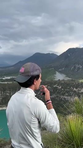 A young man wearing a white long-sleeved shirt and a baseball cap stands on a hill overlooking a valley with mountains in the background. He is holding a cigarette to his mouth and appears to be taking a drag. The sky is cloudy and overcast. There is a body of water to the left of the frame and some green foliage in the foreground.