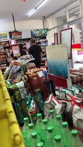 The video pans across the interior of a brightly lit convenience store. In the foreground, a row of green-capped plastic bottles, likely beverages, is visible. Behind them, various bagged snacks, including chips, are piled high in bins and on shelves. One large sign displays the word "Beras," which translates to "rice" in Indonesian. Further back, two individuals are interacting at a counter. One person, wearing a dark headscarf and possibly a mask, stands behind the counter. Another individual, wearing a hat and a dark shirt, is facing away from the camera, appearing to be a customer. The store's shelves are stocked with a variety of products, and overhead lights illuminate the space. The overall impression is that of a busy retail environment.