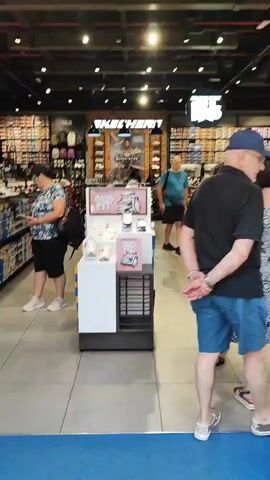 The video shows the interior of a Skechers shoe store. Multiple aisles of shoe boxes are visible, organized on shelves. In the foreground, there is a display stand with several pairs of shoes, some of which are on pedestals. There are at least four individuals visible in the scene. One woman in a patterned top and black shorts is looking at her phone. A man wearing a blue t-shirt and black shorts is walking down an aisle. Another woman in a black t-shirt and shorts is walking away from the camera. A portion of a fourth person is visible on the right side of the frame. The store appears well-lit and stocked.