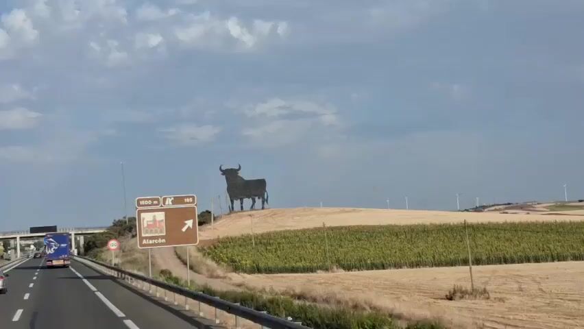 This video shows a roadside view from a moving vehicle. A large, dark silhouette of a bull stands on a hill. To the right of the bull, there is a brown road sign pointing to the right, indicating "Alarcón" at 1500 meters and highway 165. A field of green crops is visible in front of the bull, and a field of dry, light-colored crops is behind it. In the distance, several wind turbines are scattered across a rolling landscape. A truck is visible in the left lane of the highway, and a speed limit sign of 120 is on the right side of the road. The sky is partly cloudy with a mix of blue sky and white clouds. The video appears to be taken during daylight.