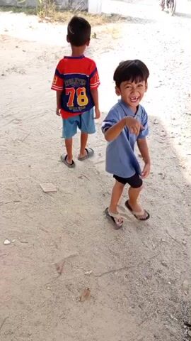 Two young boys are playing on a dusty path. The boy on the left is wearing a blue shirt and black shorts, and he is smiling and dancing. The boy on the right is wearing a red and blue shirt and blue shorts, and he is looking up and to the right. A motorcycle with a person riding it is visible in the background. The environment appears to be outdoors in a rural or suburban area with houses and vegetation visible.
