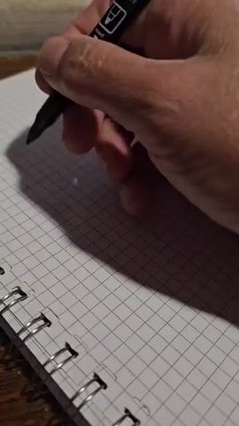 A person's hand is holding a black marker and writing on a white grid paper notebook. The writing says "I love you". The background is blurred and appears to be a wooden surface.