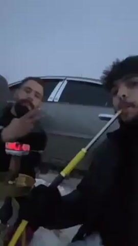 The video depicts a group of men outside in a snowy environment, possibly in the late evening or early morning due to the overcast sky and dim lighting. The men are dressed in dark clothing, and one is seen smoking from a hookah while another makes a peace sign. A silver car is parked nearby. The scene appears to be somewhat littered with empty bottles and a snack box in the snow. The background shows a few trees and what looks like a settlement on a hill. The camera movement is unsteady, suggesting it is handheld.