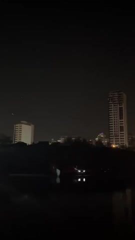 A nighttime cityscape is visible with buildings and trees silhouetted against the dark sky. In the foreground, a body of water reflects the lights from the city and two approaching headlights. The headlights appear to be from a vehicle on a road or bridge over the water. In the sky, there are three bright lights arranged in a triangular formation, with two lights positioned closely together and a third light below and between them. The lights in the sky are static in their position within the frame. The scene appears to be a still image captured from a video recording.