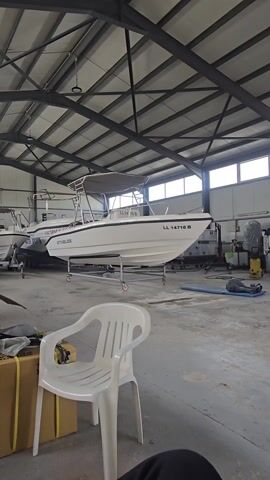 A short video shows the interior of a large warehouse with multiple white boats stored inside. The boats are placed on dollies, allowing for easy movement. The camera pans slightly to the left, revealing more of the warehouse's interior. A white plastic chair sits in the foreground, with various objects, including a cardboard box and some other items, placed around it. The warehouse has a high ceiling with visible metal beams. Natural light filters in through the windows, illuminating the scene. No people are visible in the frame. The environment appears to be a storage or maintenance facility for boats.
